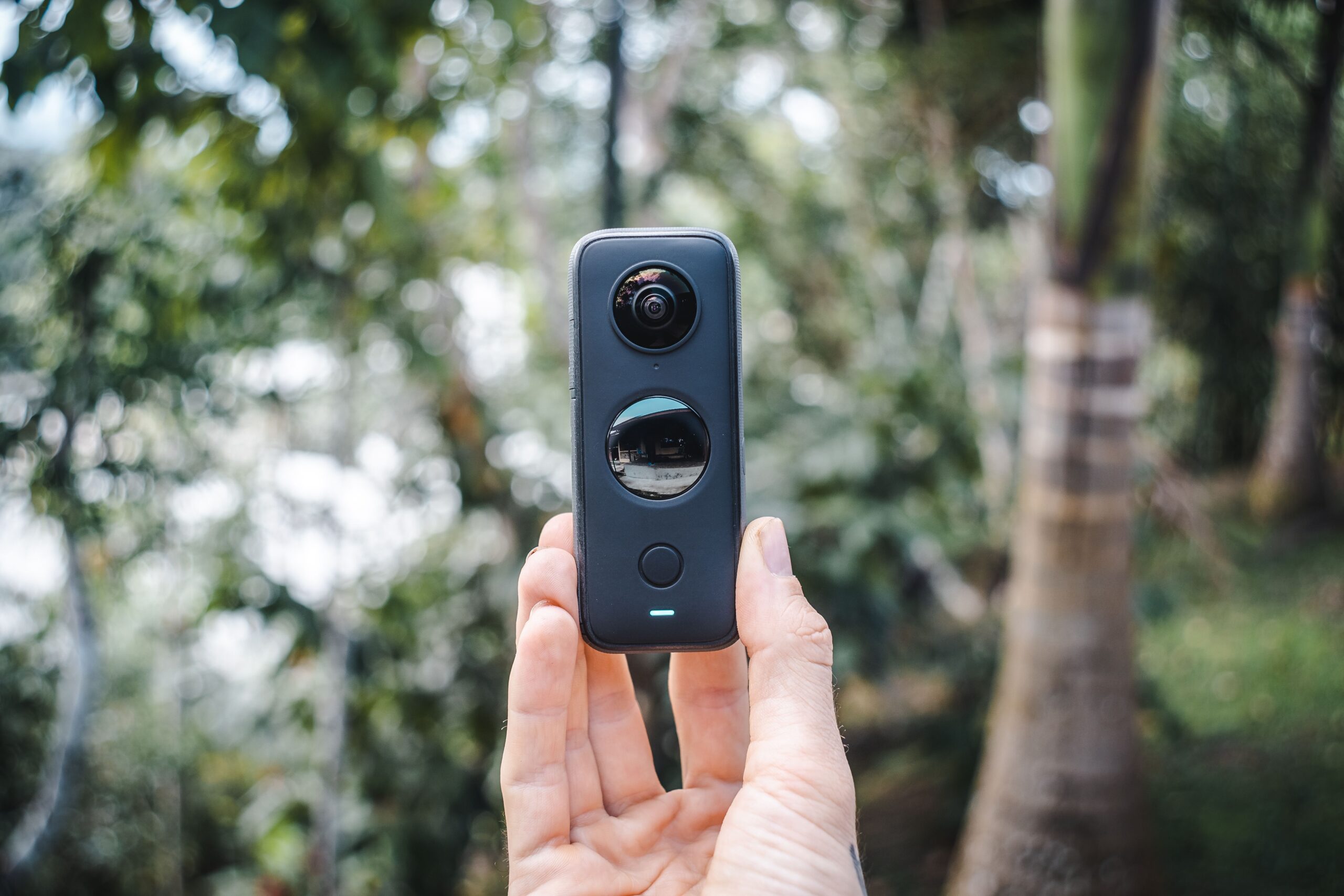 Close-up of a hand holding a compact 360 camera outdoors with greenery.